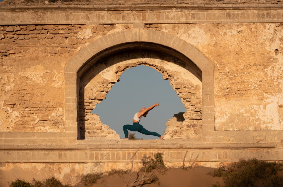 Manon Descroix en posture du guerrier 1 (Virabhadrasana) encadrée par une arche de pierre en plein air